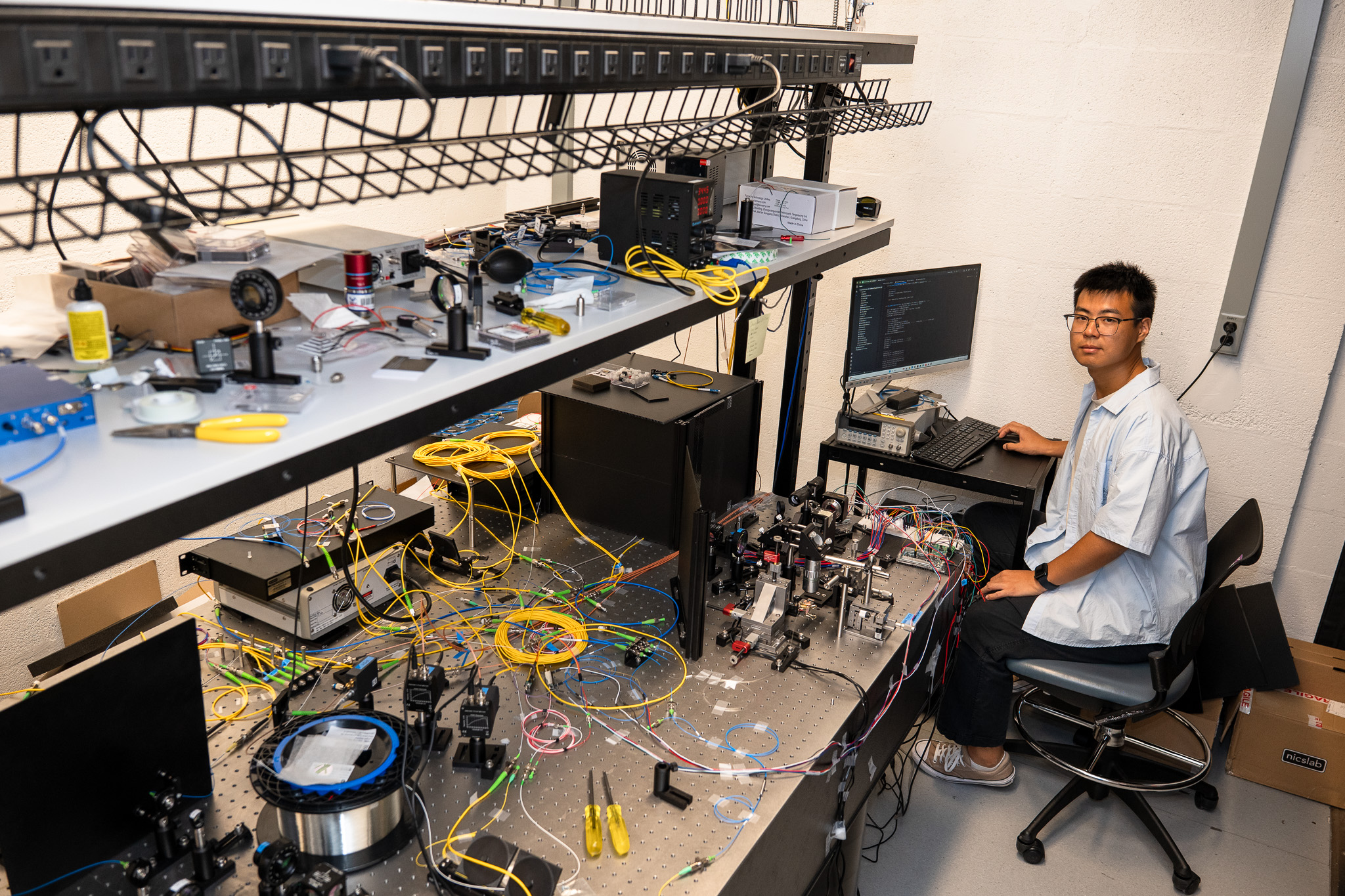 A scientist sits at a computer next to a testbed of wires.